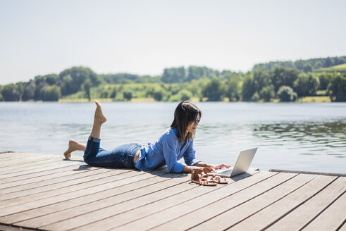 Reife Frau bei der Arbeit an einem See, mit Laptop auf einem Steg - MOEF01771