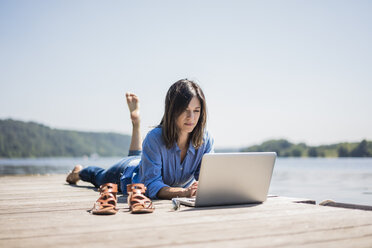 Reife Frau bei der Arbeit an einem See, mit Laptop auf einem Steg - MOEF01772