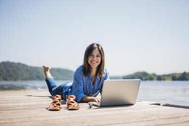 Reife Frau bei der Arbeit an einem See, mit Laptop auf einem Steg - MOEF01773