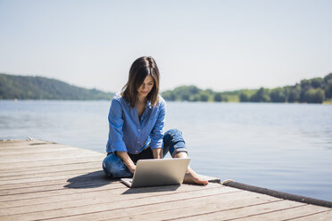 Mature woman working at a lake, using laptop on a jetty - MOEF01775