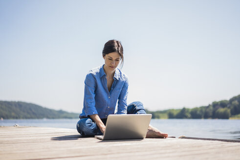 Mature woman working at a lake, using laptop on a jetty - MOEF01776