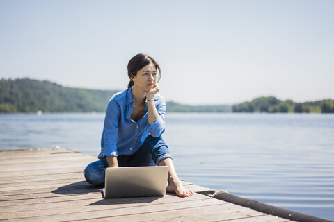 Reife Frau bei der Arbeit an einem See, mit Laptop auf einem Steg, lizenzfreies Stockfoto