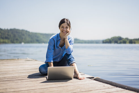 Reife Frau bei der Arbeit an einem See, mit Laptop auf einem Steg - MOEF01778