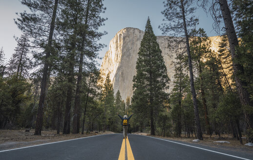 USA, Kalifornien, Yosemite National Park, Mann mit erhobenen Armen auf der Straße mit El Capitan im Hintergrund - KKAF03035