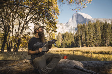 USA, Kalifornien, bärtiger Mann mit einer Landkarte auf einem Baumstamm im Yosemite-Nationalpark sitzend - KKAF03057