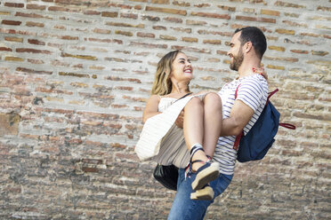 Happy man with backpack carrying girlfriend at brick wall - JSMF00604