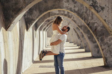 Spain, Andalusia, Malaga, happy man lifting up girlfriend under an archway in the city - JSMF00629