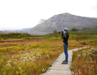 Seitenansicht eines Wanderers, der mit der Kamera fotografiert, während er auf der Uferpromenade vor den Bergen steht - CAVF57925