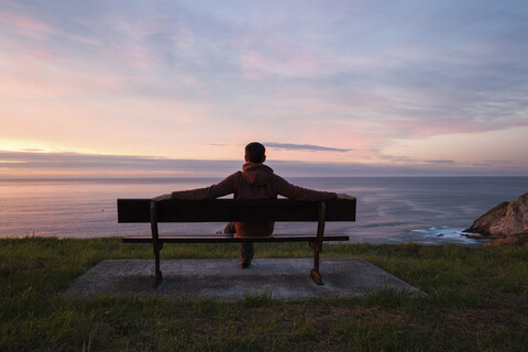Mann in voller Länge mit Blick auf das Meer, während er auf einer Bank sitzt, gegen den bewölkten Himmel bei Sonnenuntergang, lizenzfreies Stockfoto