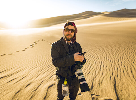 Porträt eines Wanderers, der sein Smartphone in der Hand hält, während er im Great Sand Dunes National Park steht, lizenzfreies Stockfoto