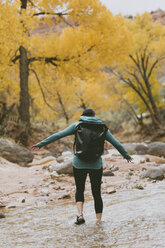 Rear view of hiker with backpack wading in stream during autumn at forest - CAVF57948
