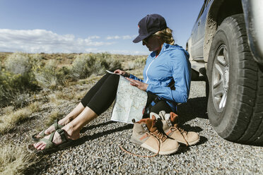 Wanderin in voller Länge, die eine Karte liest, während sie neben einem Auto auf der Straße sitzt - CAVF57953