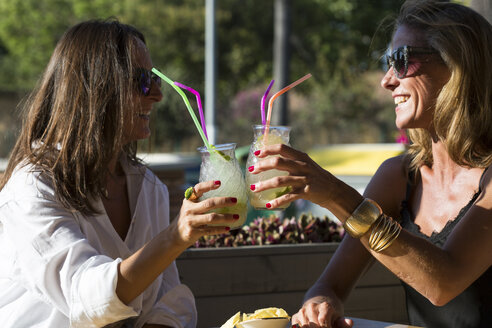 Fröhliche Freundinnen sitzen im Freien und stoßen mit Cocktailgläsern an - ERRF00230