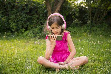 Porträt eines Mädchens, das auf einer Wiese sitzt und ein Smartphone und rosa Kopfhörer benutzt - LVF07577