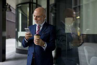 Älterer Geschäftsmann mit Mobiltelefon und Kaffee zum Mitnehmen an der Glasfront - MAUF01807