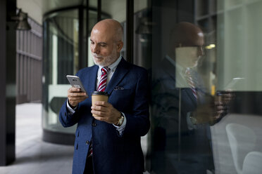 Senior businessman with cell phone and takeaway coffee at glass front - MAUF01807