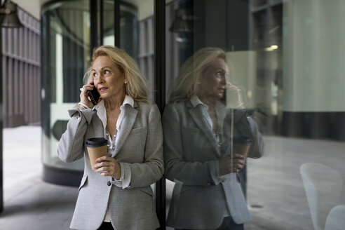 Ältere Geschäftsfrau lehnt an einer Glasfront und spricht mit einem Handy - MAUF01809