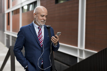 Senior businessman with cell phone and earphones outdoors - MAUF01815