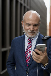 Senior businessman with cell phone and earphones outdoors - MAUF01816