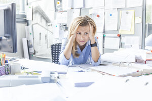 Verzweifelte Frau sitzt am Schreibtisch im Büro, umgeben von Papierkram - TCF06045