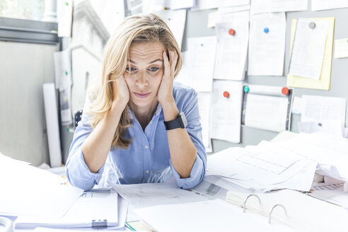 Stressed woman sitting at desk in office surrounded by paperwork - TCF06046