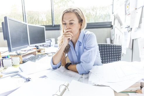 Pensive woman sitting at desk in office surrounded by paperwork - TCF06050