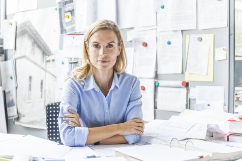 Porträt einer selbstbewussten Frau, die an einem Schreibtisch im Büro sitzt, umgeben von Papierkram - TCF06057