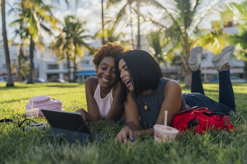Zwei glückliche Freundinnen entspannen sich in einem Park und benutzen ein Tablet, lizenzfreies Stockfoto