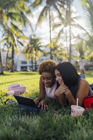 Zwei glückliche Freundinnen entspannen sich in einem Park und benutzen ein Tablet, lizenzfreies Stockfoto