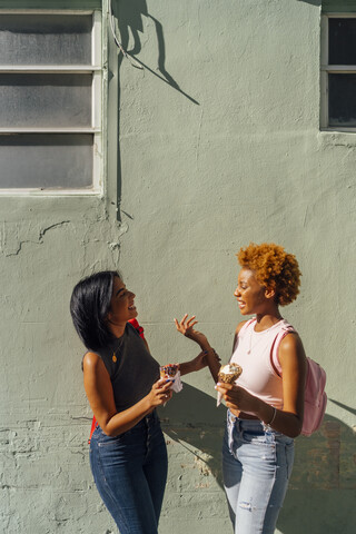 Two happy female friends with ice cream cones talking at a house wall stock photo