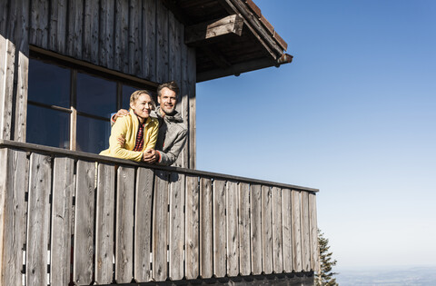 Paar lehnt auf dem Balkon einer Berghütte, die Arme um sich gelegt, lizenzfreies Stockfoto