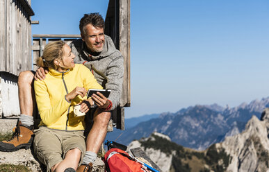 Hiking couple sitting in front of mountain hut, taking a break, looking at smartphone - UUF15972