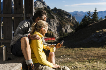 Wanderpaar vor einer Berghütte sitzend, eine Pause einlegend, auf das Smartphone schauend - UUF15973