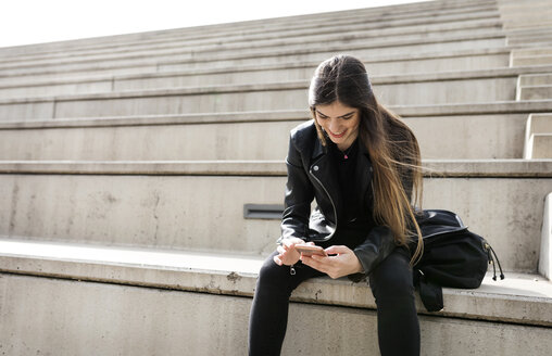 Lächelnde junge Frau sitzt auf einer Treppe und benutzt ein Mobiltelefon - VABF01940