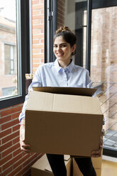 Porträt einer lächelnden jungen Frau, die einen Karton in einer neuen Wohnung am Fenster trägt - VABF02017