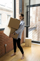 Porträt einer lächelnden jungen Frau, die einen Karton in einer neuen Wohnung am Fenster trägt - VABF02019