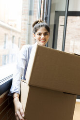 Porträt einer lächelnden jungen Frau, die einen Karton in einer neuen Wohnung am Fenster trägt - VABF02020