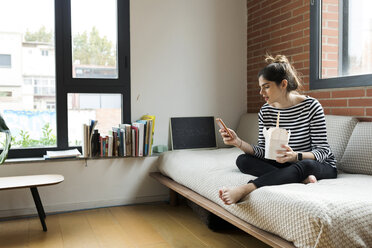 Junge Frau sitzt zu Hause auf der Couch, isst asiatisches Essen zum Mitnehmen und benutzt ein Mobiltelefon - VABF02022