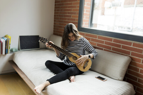 Junge Frau sitzt auf der Couch zu Hause und spielt Gitarre, lizenzfreies Stockfoto