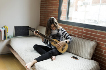 Young woman sitting on couch at home playing guitar - VABF02027