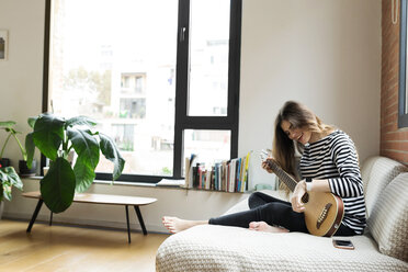 Happy young woman sitting on couch at home playing guitar - VABF02028