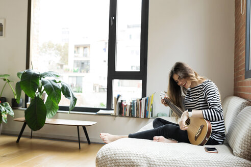 Glückliche junge Frau sitzt auf der Couch zu Hause und spielt Gitarre - VABF02028