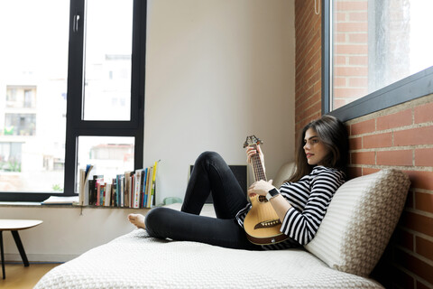 Junge Frau sitzt auf der Couch zu Hause und spielt Gitarre, lizenzfreies Stockfoto