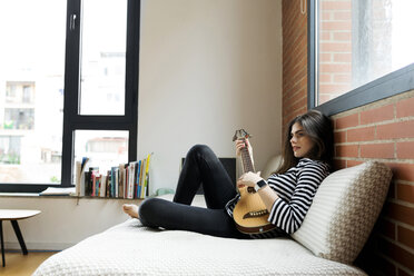 Young woman sitting on couch at home playing guitar - VABF02029