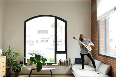 Glückliche junge Frau, die zu Hause auf der Couch steht und Gitarre spielt - VABF02031