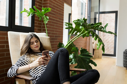 Junge Frau sitzt in einem Sessel zu Hause und benutzt ein Mobiltelefon, lizenzfreies Stockfoto