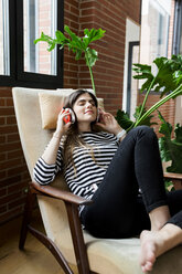 Young woman sitting in armchair at home listening to music with headphones - VABF02034