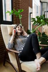 Happy young woman sitting in armchair at home listening to music with headphones - VABF02035