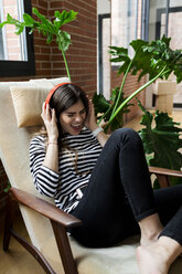 Excited young woman sitting in armchair at home listening to music with headphones - VABF02036