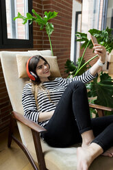 Junge Frau, die zu Hause im Sessel sitzt und mit Kopfhörern Musik hört und ein Selfie macht - VABF02037